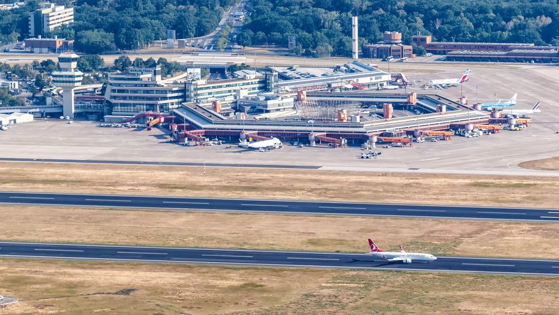Das Bild zeigt eine Luftaufnahme eines Flughafens mit einem Flugzeug, das von der Startbahn abhebt. Im Vordergrund ist die Startbahn zu sehen, die von einer Reihe von Gebäuden und Hangars flankiert wird. Im Hintergrund ist eine große Fläche mit Grasland zu sehen.